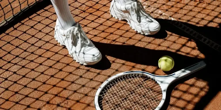 Tennis players legs with racket and ball on clay court under bright sun and shadow patterns
