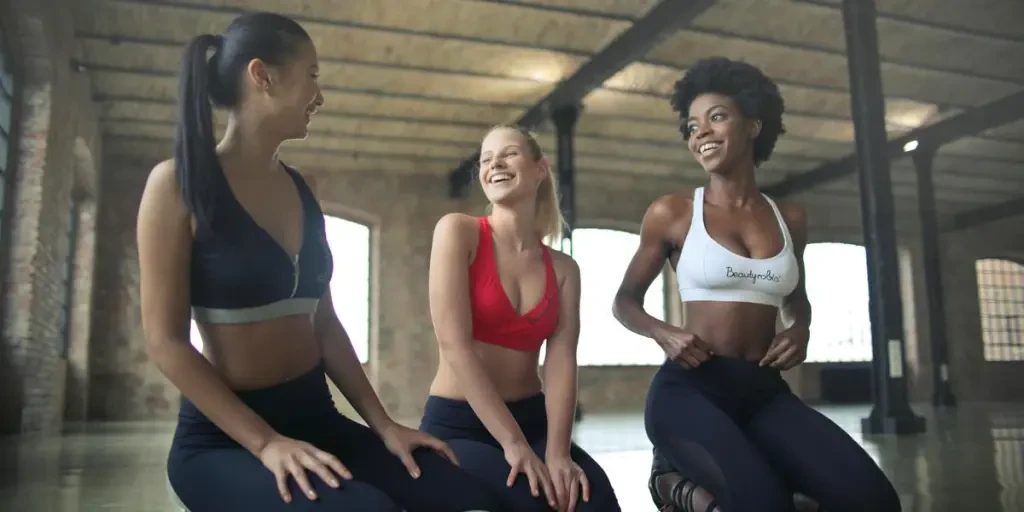 Three women in activewear laughing and exercising together indoors in a gym setting