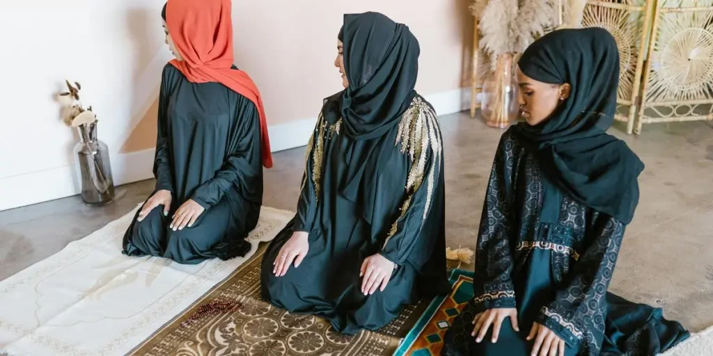 Three women in traditional attire and hijabs praying indoors on beautiful rugs