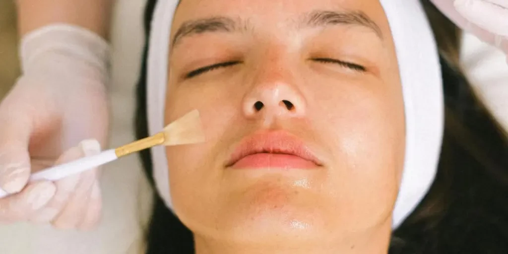 Top view skilled female cosmetologist in uniform and gloves applying essential oil on young woman face skin while working in contemporary spa center