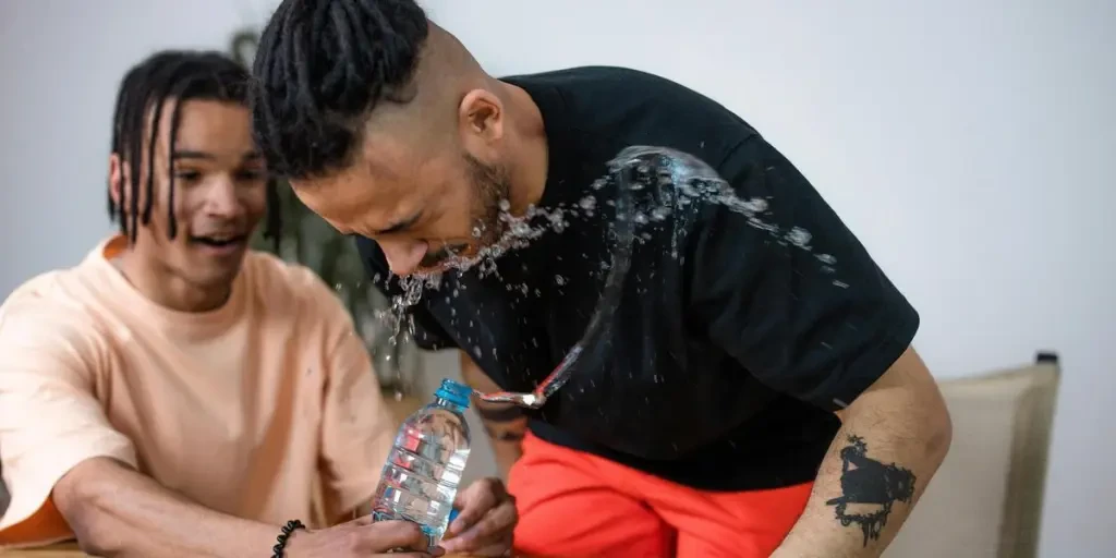 Two adult friends laughing during a playful water splash prank at a wooden table indoors