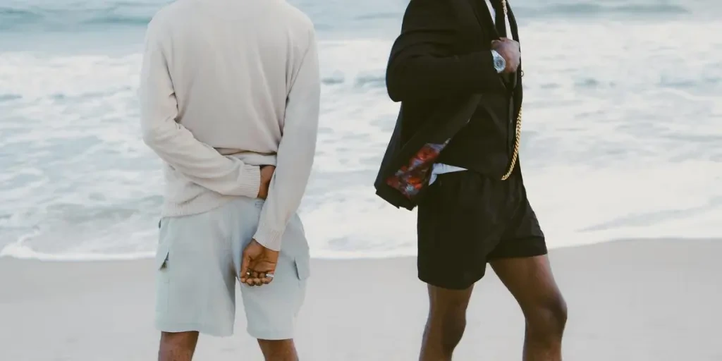 Two fashionable men standing on a beach with modern outfits and vibrant socks, facing the ocean