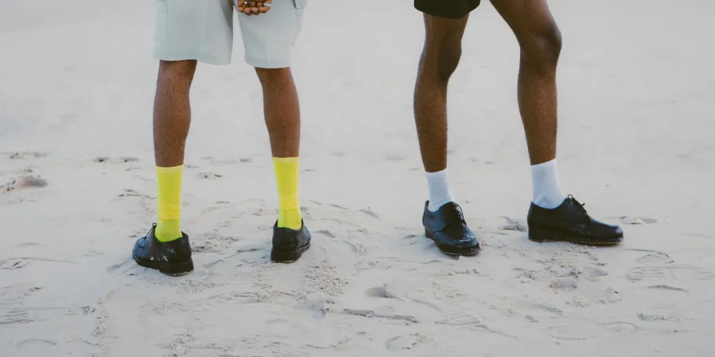 Two fashionable men standing on a beach with modern outfits and vibrant socks, facing the ocean