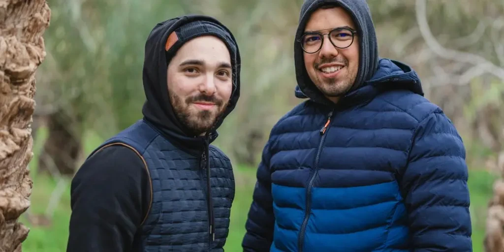 Two men warmly dressed in hooded jackets smile outdoors, showcasing winter fashion