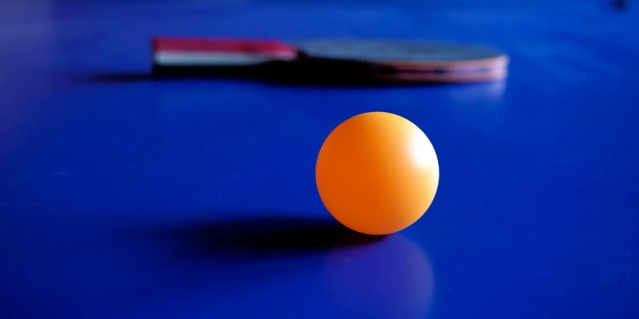 Vibrant orange ping pong ball with a table tennis racket on a blue table, emphasizing sport and leisure