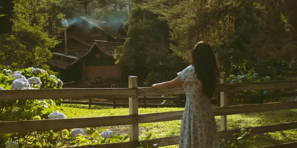 Woman in a floral dress views a serene countryside house from a wooden fence 