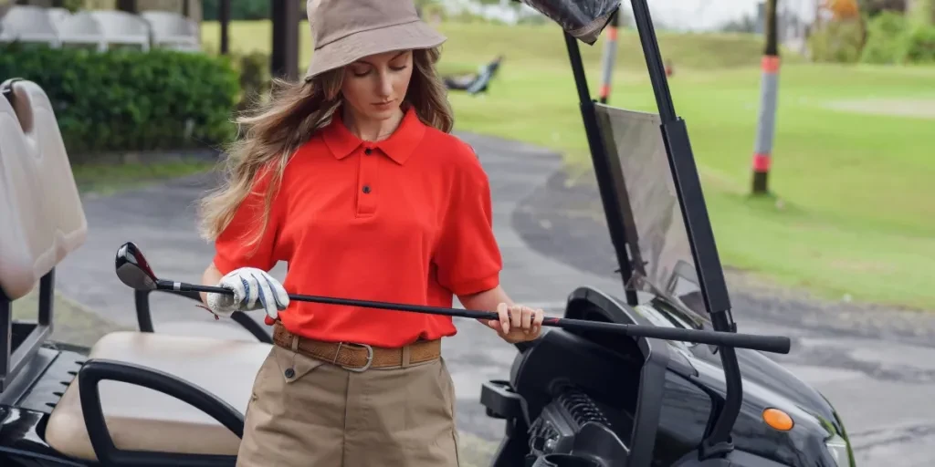 Woman in red and khaki golfing outfit standing by a cart, holding a golf club on a sunny day