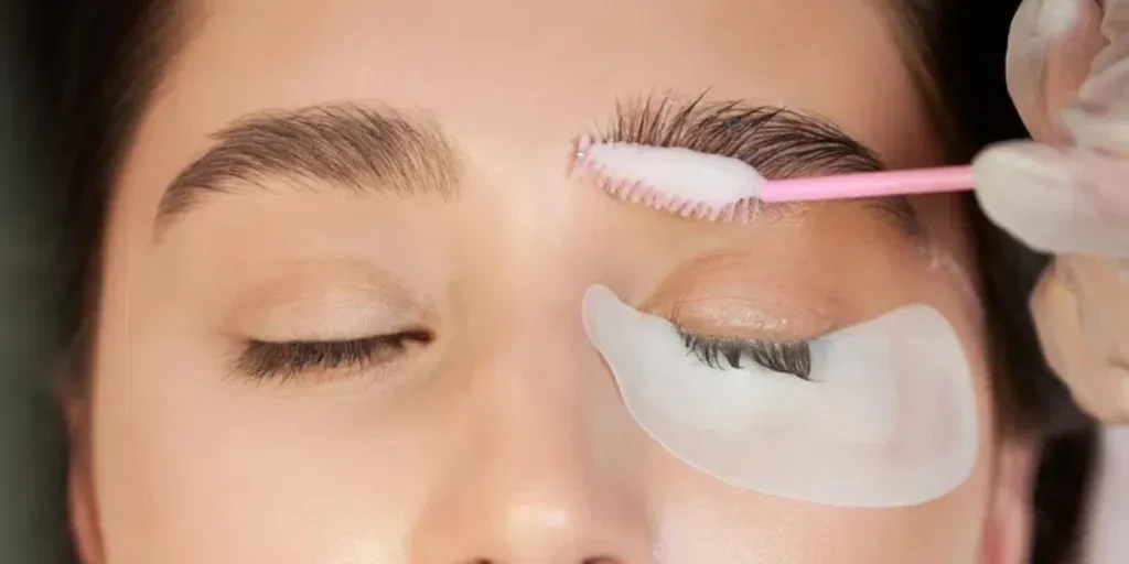 Woman with eye patch under lower eyelid keeping eyes closed while beautician applying cleansing mousse on eyebrows