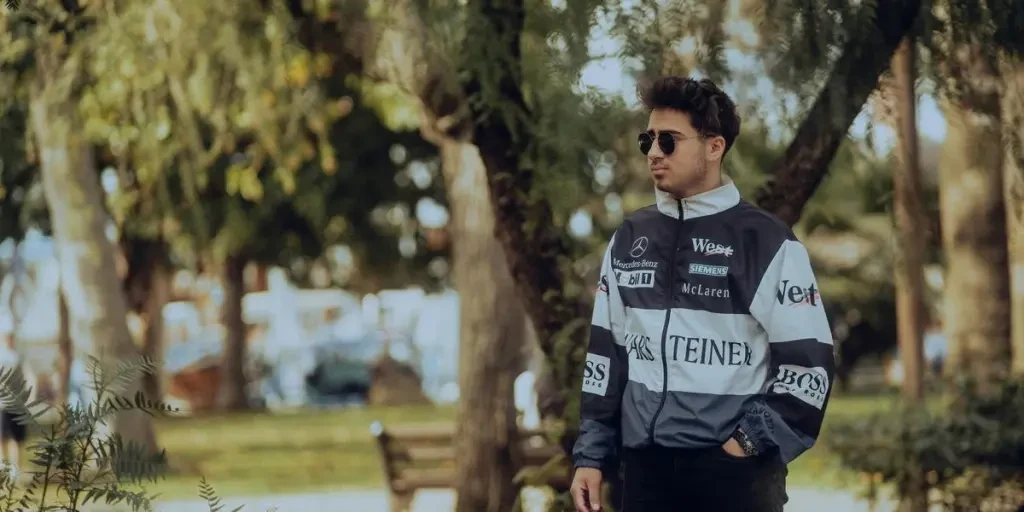 Young adult male in sunglasses and racing jacket standing in a park setting, exuding cool style