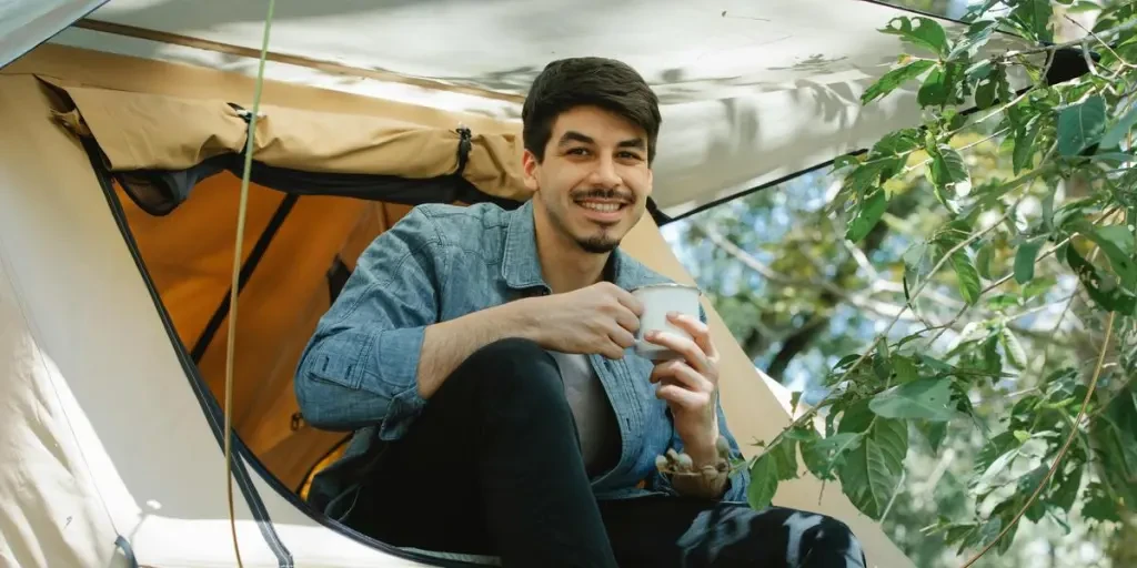 Young man enjoying coffee at a campsite, smiling and relaxed outdoor setting