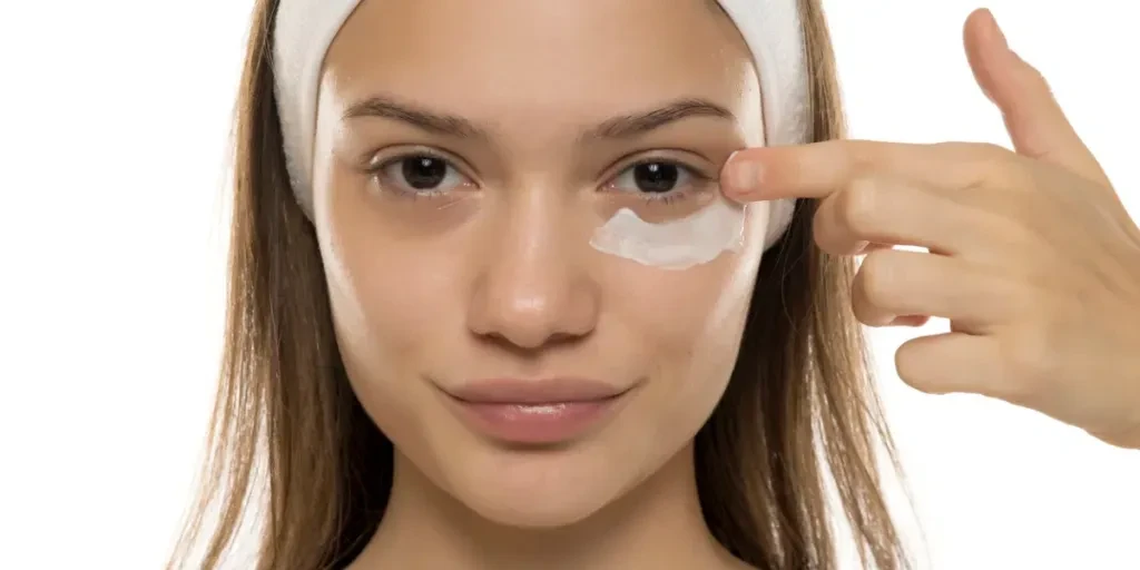 Young smiling woman applying cosmetic cream under her eyes on a white background