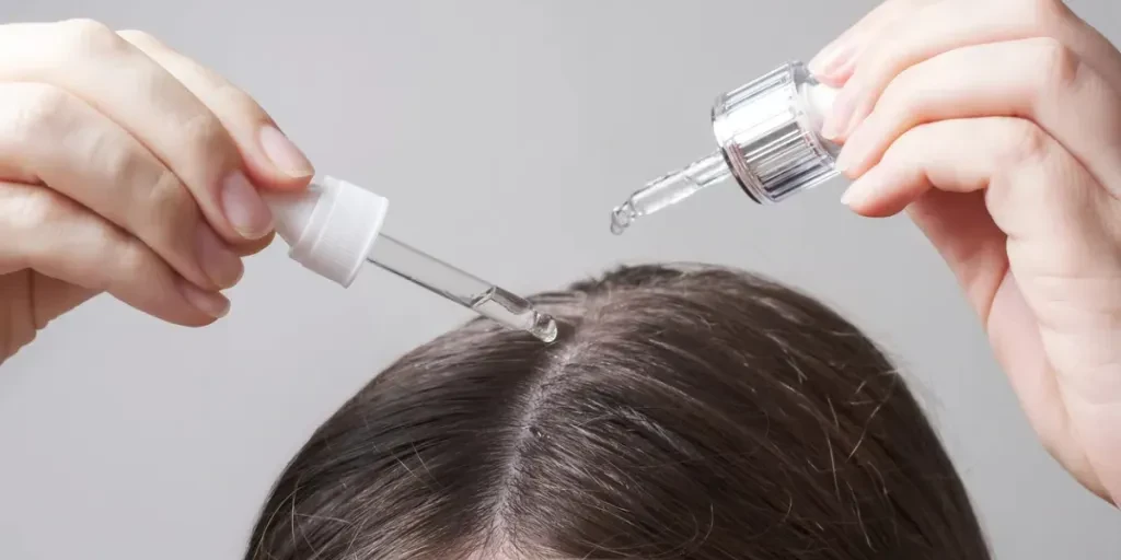 Young woman applying oil to her hair with a pipette on a gray background close up