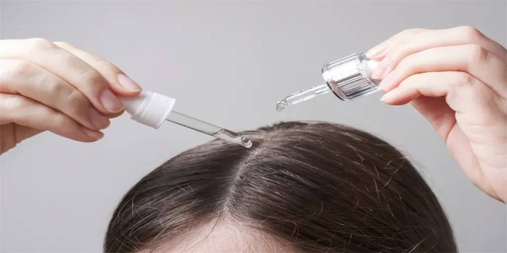 Young woman applying oil to her hair with a pipette on a gray background close up