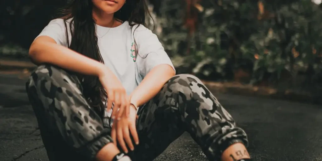 Young woman sitting on pavement in a casual, stylish pose with camouflage pants and sneakers