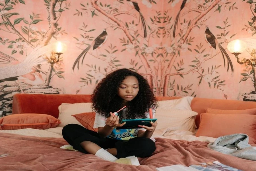 A girl applying lipstick with a wallpapered wall behind her