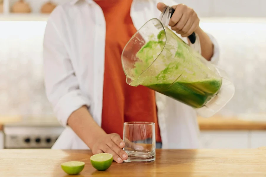 Close-up of a person pouring a vibrant green smoothie into a glass with limes on the wooden table