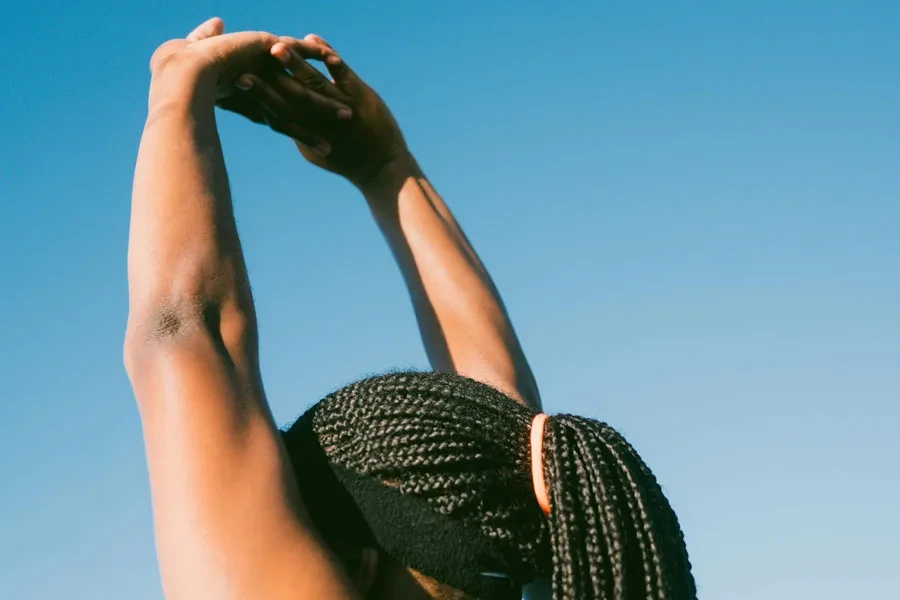 Person stretching outdoors with braided hair against a clear blue sky for a healthy lifestyle concept