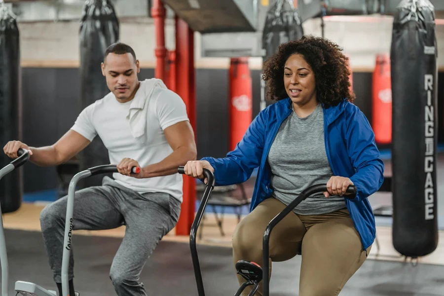 Plus-sized African American female in sportswear exercising on a stationary bicycle near fit muscular male coach in the modern gym