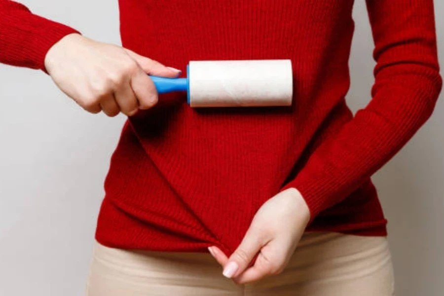 Mano de mujer usando un rodillo adhesivo para limpiar telas (cuello alto de lana roja) de polvo, pelo, pelusa y pelusa.