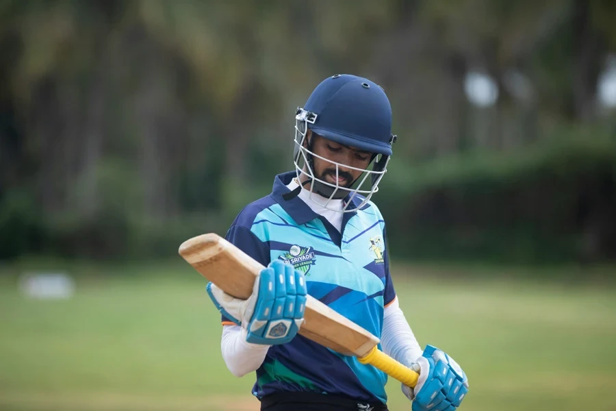 A cricketer in gear, holding a bat, prepares for a match outdoors