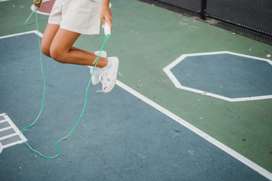 A girl playfully jumping rope on a painted outdoor playground surface, wearing casual shorts and sneakers