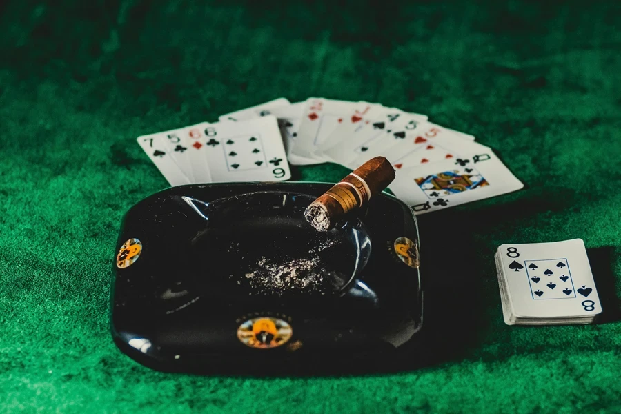 A poker table with playing cards, a lit cigar, and an ashtray on a green felt surface