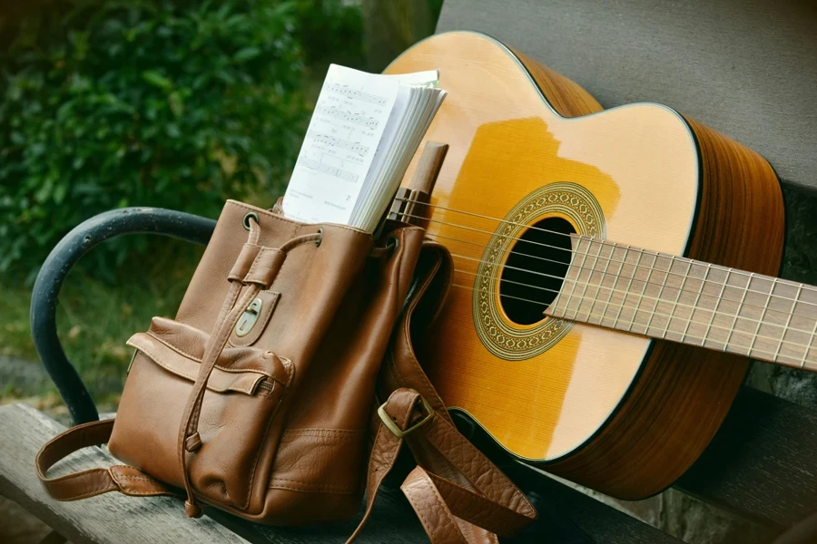 A serene outdoor scene of an acoustic guitar and a brown backpack on a wooden bench, perfect for music lovers