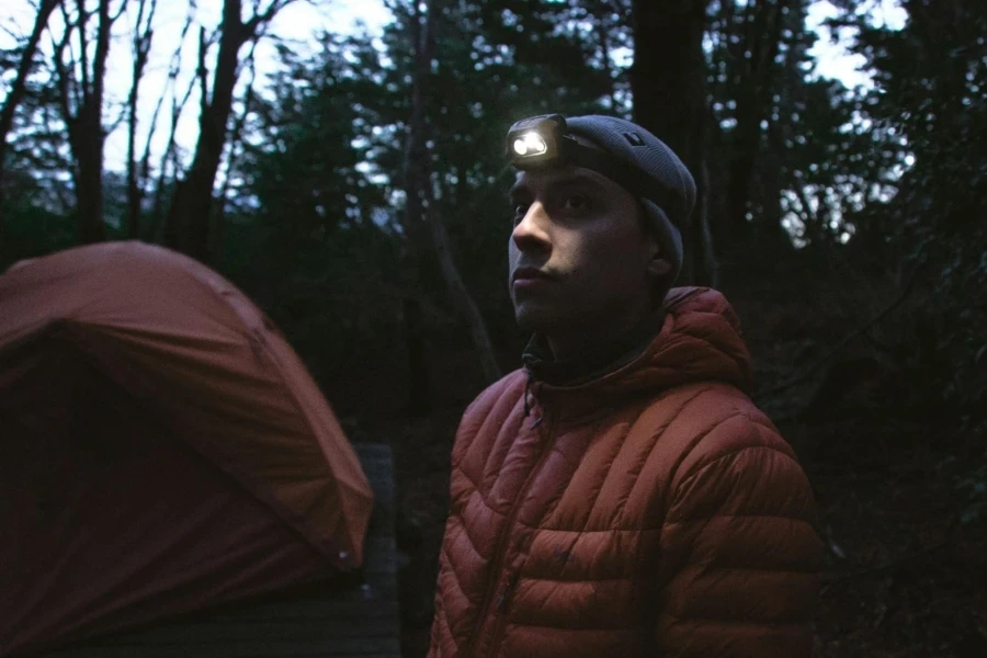 Man in a Winter Jacket and a Headlamp next to a Tent in the Forest