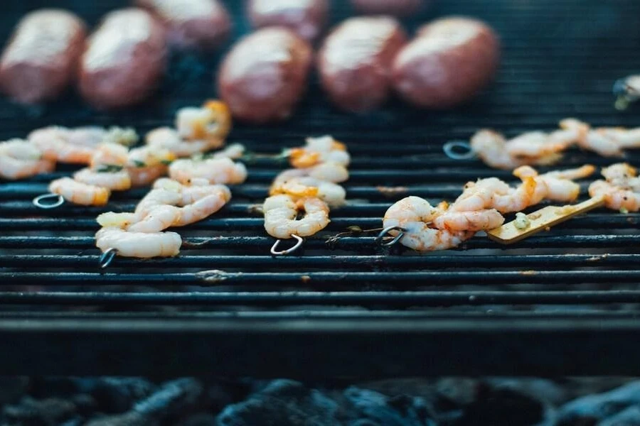 Several stainless steel skewers grilling small shrimp on barbecue