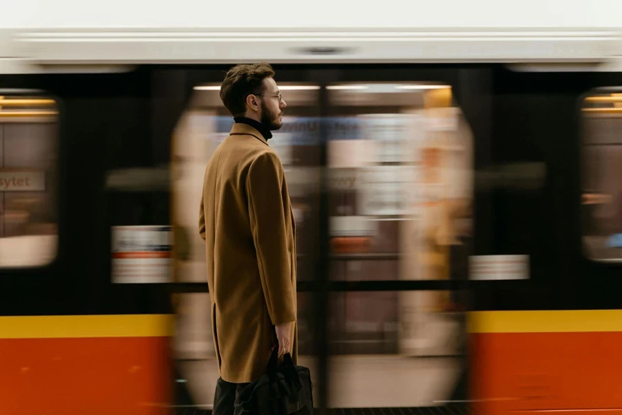 A Man in Brown Coat Standing on a Train Station Near a Moving Train