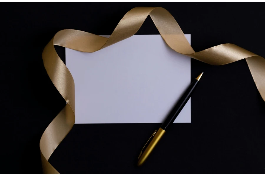 A gold pen, a white piece of paper and a gold ribbon lie on a white background.
