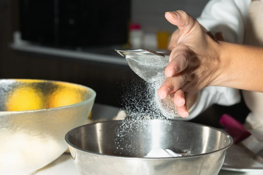 Baker using small mesh sifter over stainless steel mixing bowl