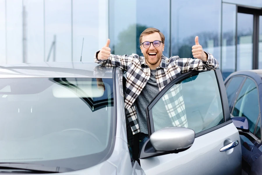 Cheerful guy standing near auto, gesturing thumbs up, buying vehicle in dealership center