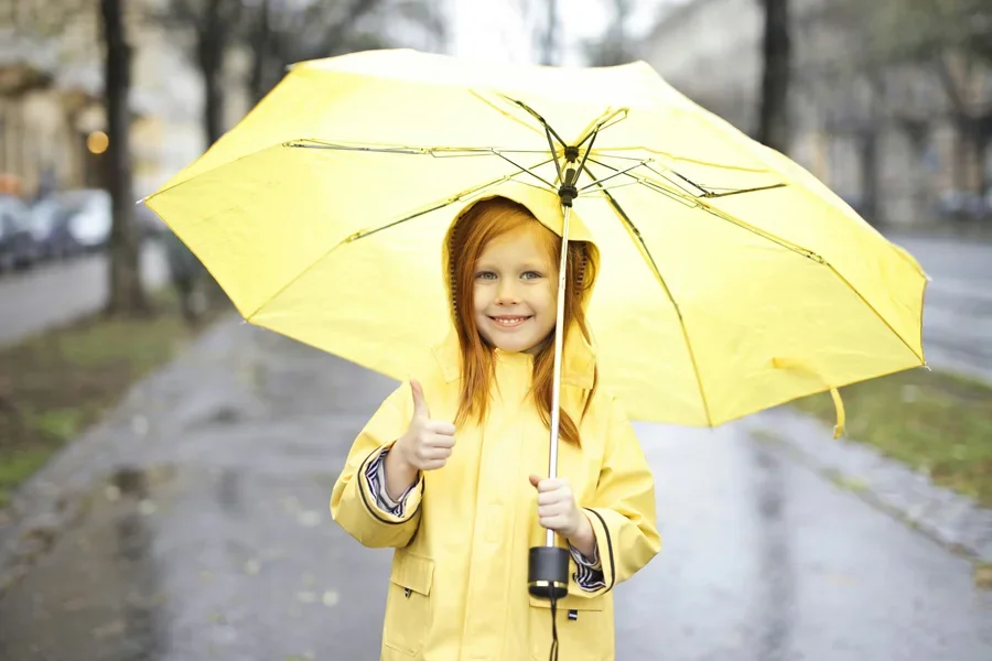 Niña sonriendo bajo un paraguas amarillo en una calle lluviosa, mostrando un pulgar hacia arriba