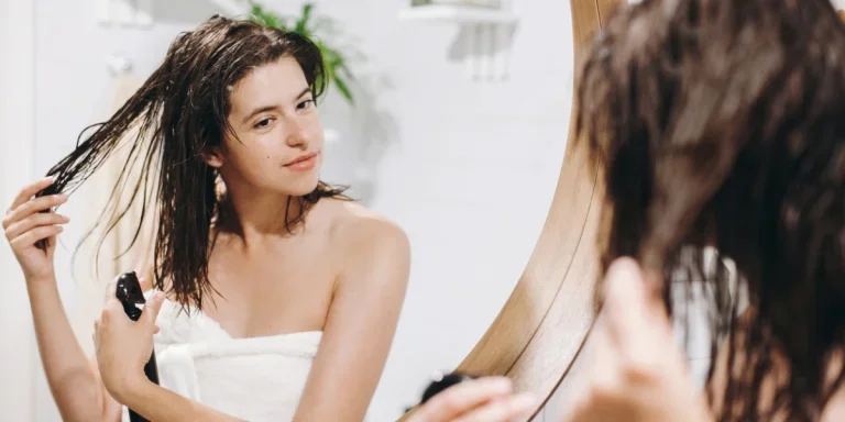 Young happy woman in white towel applying conditioner mask on hair in bathroom, mirror reflection.