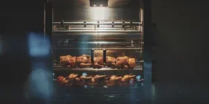 Gambar dari dekat menunjukkan ayam panggang yang berputar dalam oven rotisserie logam mengilap yang terang benderang dan berpintu kaca.