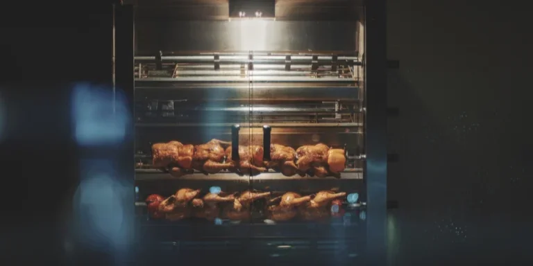 Крупным планом показаны жареные цыплята, вращающиеся в ярко освещенной, отполированной металлической вертеле со стеклянной дверцей.