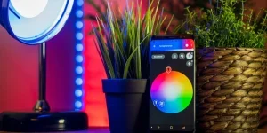 A smart home setup features a black digital clock displaying. Both devices are positioned on a wooden surface in a softly lit indoor environment.