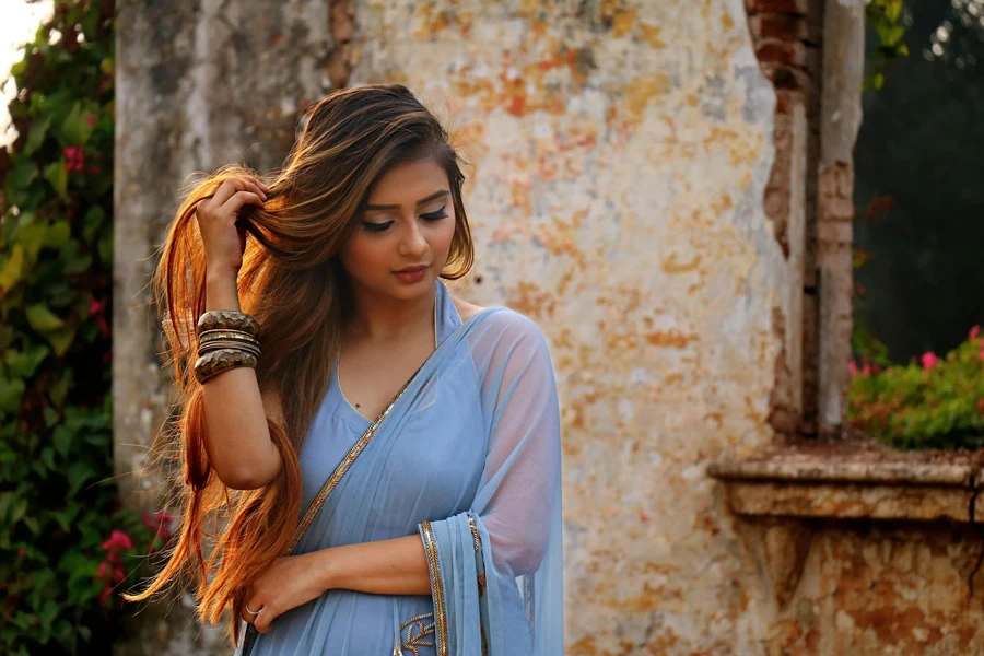 A woman in a blue saree poses gracefully outdoors by a rustic wall