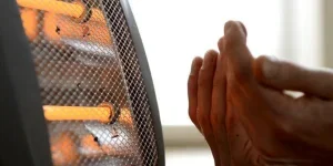 An individual sits comfortably on a sofa, reading a book while a small electric fan heater, positioned on the floor, provides warmth