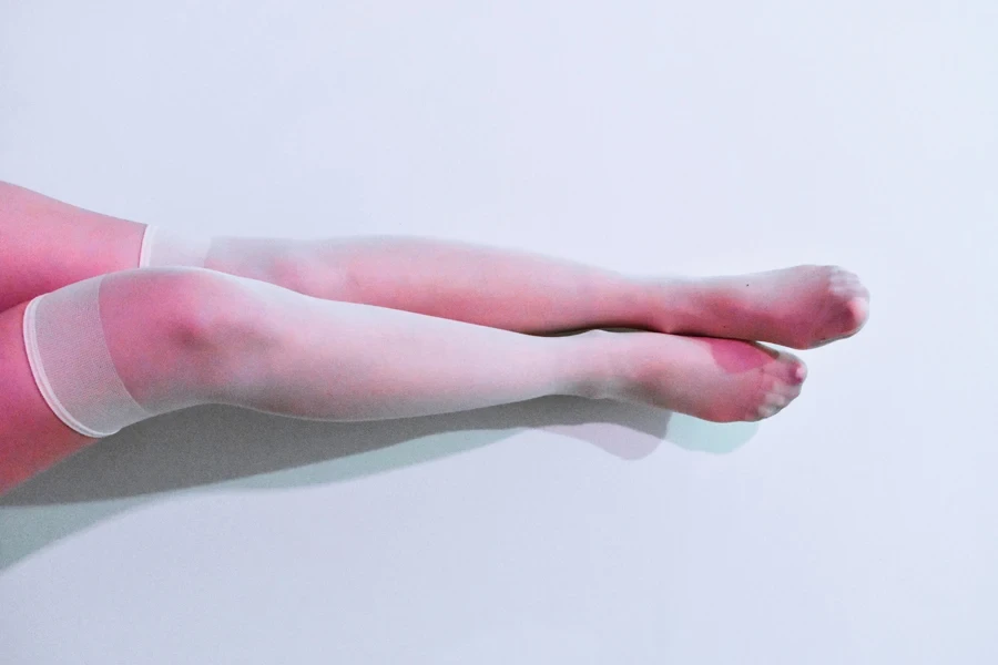 Close-up of female legs in white stockings on a pastel pink backdrop, creating a modern and minimalist aesthetic