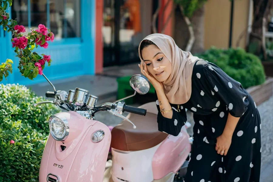 Modisch gekleidete Frau mit Kopftuch bewundert ihr Spiegelbild im Spiegel eines Motorrollers
