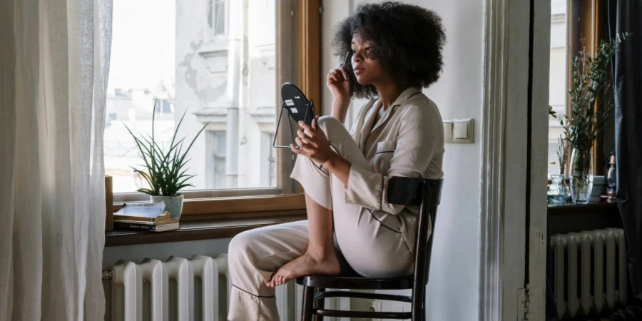 Woman in pajamas applying skincare with mirror, enjoying a cozy morning by the window