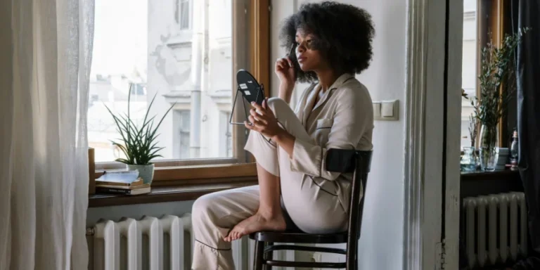 Mujer en pijama aplicándose productos para el cuidado de la piel frente a un espejo, disfrutando de una mañana acogedora junto a la ventana.