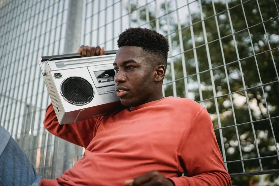 Young pondering African American male listening to radio from old tape recorder while looking forward near grid fence in town