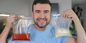 A handsome bearded man in his early forties holding one plastic bag with spaghetti and one plastic bag with tomato sauce