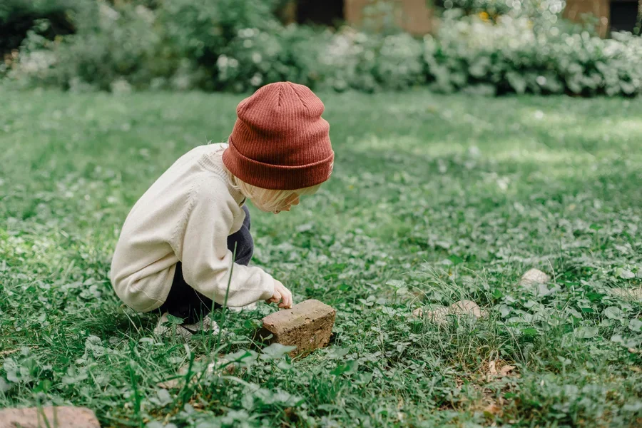 Niño con sombrero rojo y suéter blanco agachado sobre un ladrillo en el césped