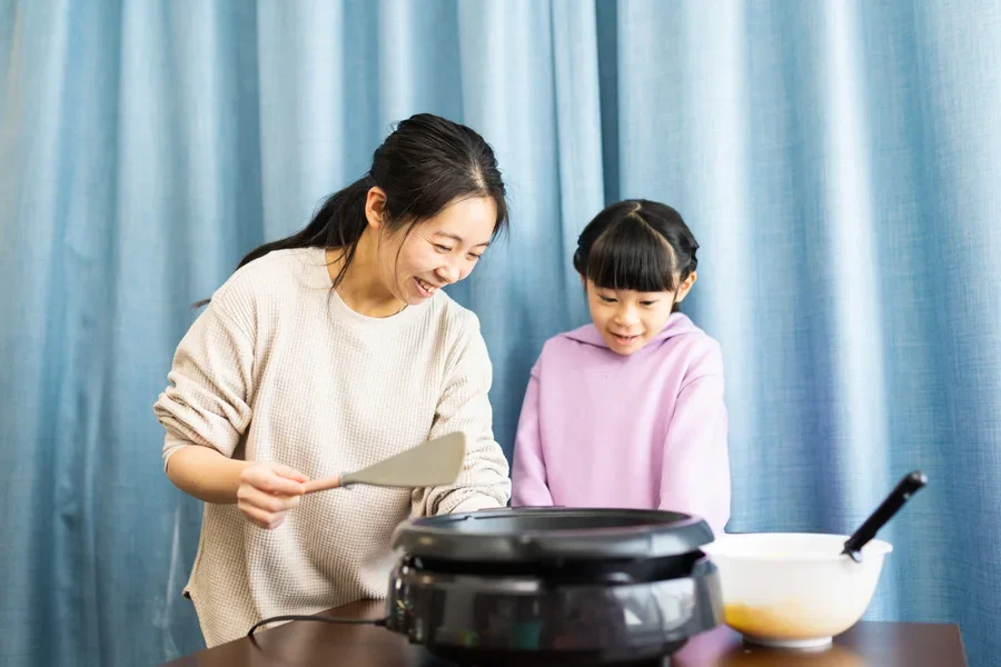 Мать и дочь готовят блины с помощью электрической блинницы.