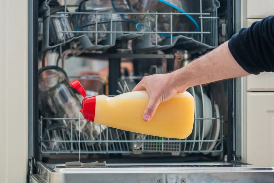 Seseorang menuangkan cairan pembilas berwarna kuning ke dalam dispenser di dalam mesin pencuci piring
