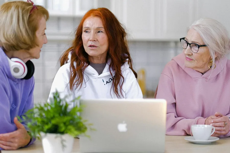 Tres mujeres mayores charlando con una computadora portátil y una taza de té, adoptando la tecnología en interiores.